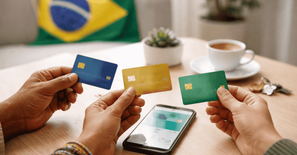 Imagem que representa a escolha e utilização de cartões bancários no contexto financeiro brasileiro. Em primeiro plano, duas pessoas seguram três cartões bancários de cores diferentes — azul, dourado e verde — enquanto os comparam, possivelmente avaliando opções de pagamento ou benefícios de cada cartão. Sobre a mesa encontra-se um smartphone com uma aplicação financeira aberta, sugerindo gestão digital de cartões ou comparação de serviços bancários. No fundo da cena aparece uma chávena de café, um pequeno vaso com planta e um conjunto de chaves, criando um ambiente doméstico. Ao fundo também é visível uma bandeira do Brasil, indicando o contexto do sistema financeiro ou do mercado de cartões no país.